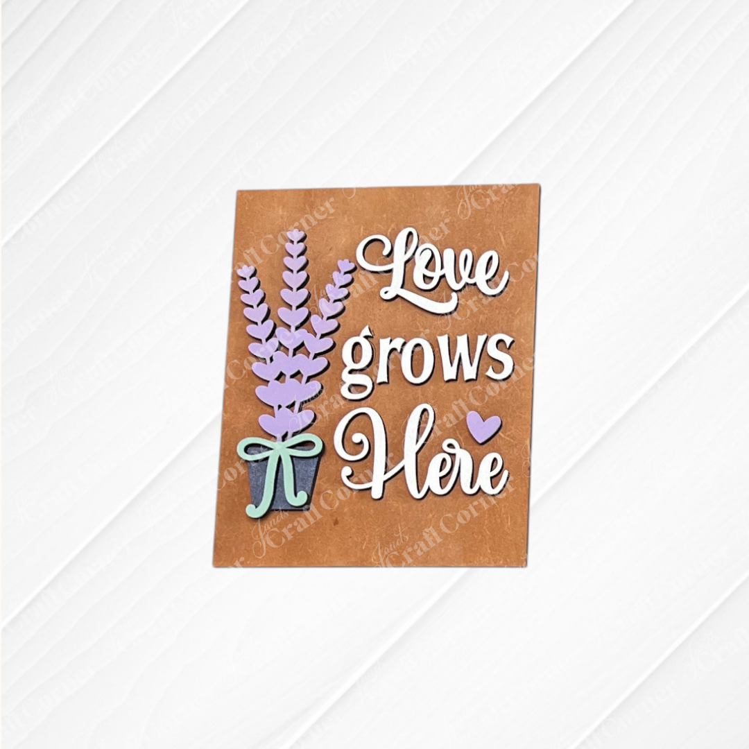 Janet's Craft Corner’s Lavender Switchable Farmhouse Sign features a brown card with a potted lavender, "Love grows here" in white script, purple heart, and blue bow—perfect DIY decor for tiered trays or small spaces. Set of 1.