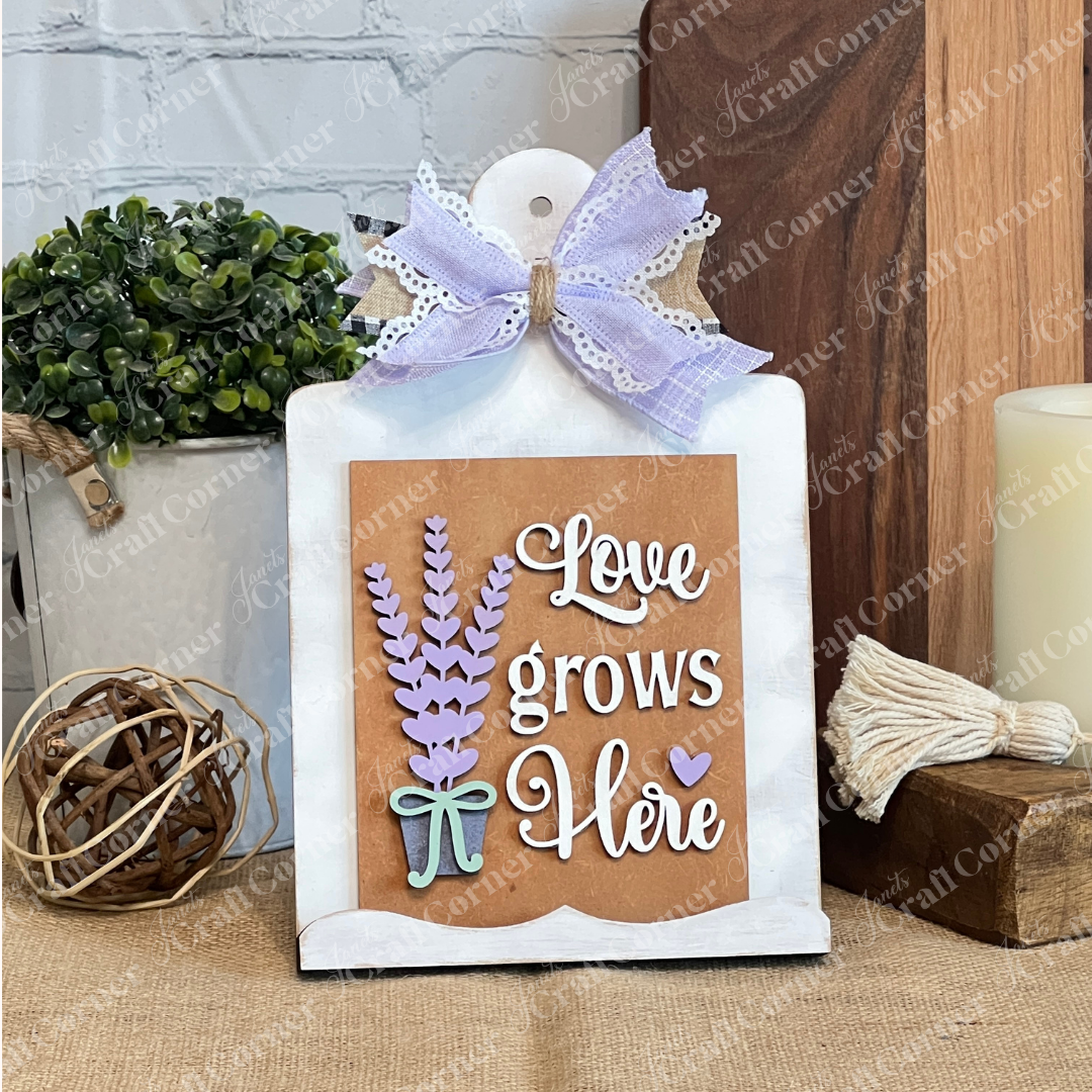 The Lavender Switchable Farmhouse Signs DIY Craft Kit by Janet's Craft Corner features a sign with a bow and "Love grows here," perfect for a white stand, rustic tiered tray décor, and a small potted plant for charming farmhouse style.