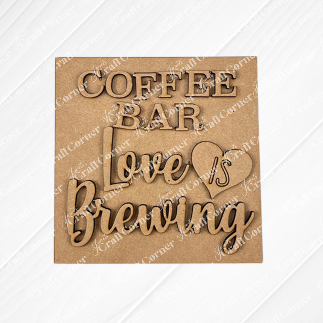 A wooden sign with layered text reads "COFFEE BAR Love is Brewing," with "is" in a heart, on a white textured background—perfect for Janet's Craft Corner Coffee Bar Swappable Squares DIY seasonal home decor craft kit.