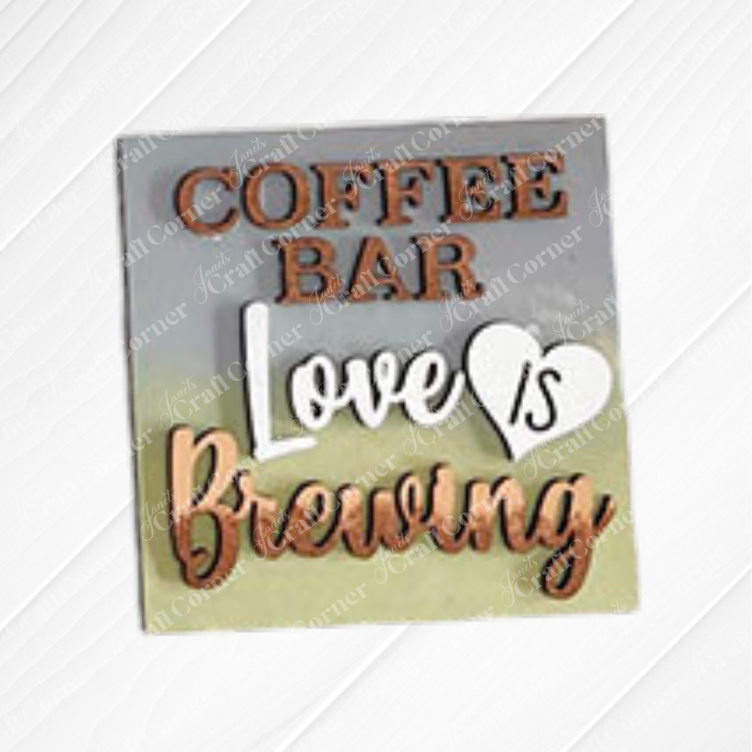 A small blue sign from Janet’s Craft Corner’s Coffee Bar Swappable Squares collection sits on a white wooden surface, reading "COFFEE BAR Love IS Brewing" with "IS" inside a white heart—perfect for cozy DIY home decor.