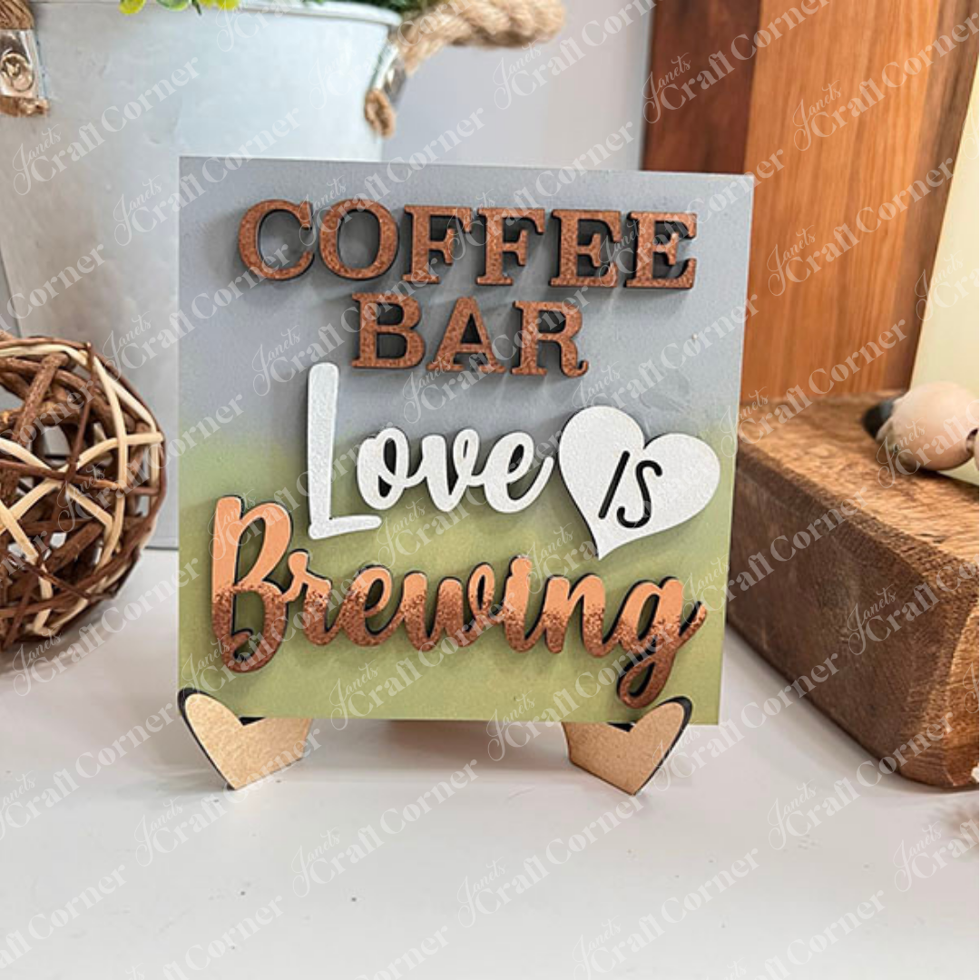 A small blue sign from Janet’s Craft Corner’s Coffee Bar Swappable Squares collection sits on a white surface against a white brick wall, reading "COFFEE BAR Love IS Brewing" with "IS" inside a white heart—perfect for cozy DIY home decor.