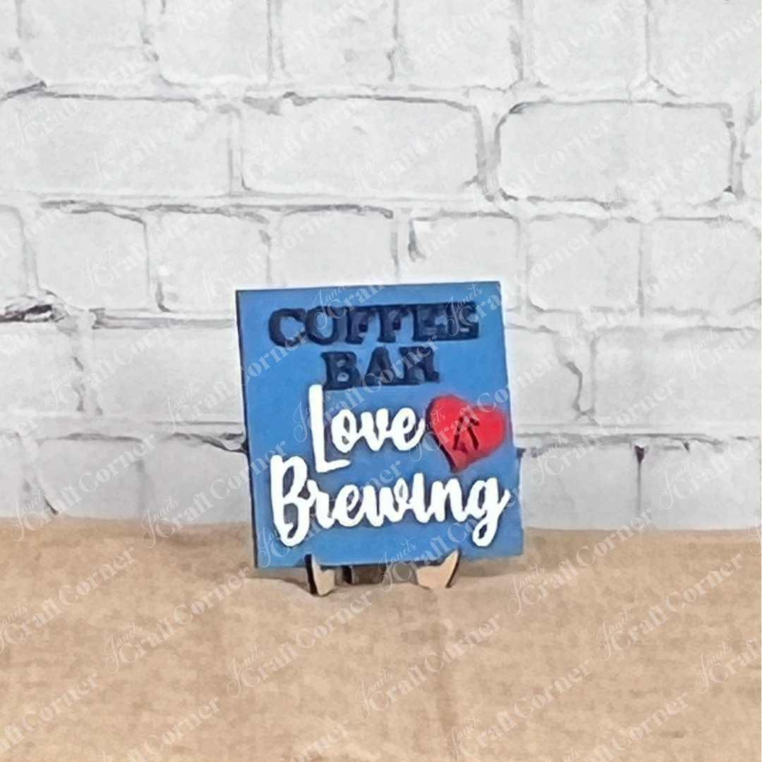 A small blue sign from Janet’s Craft Corner’s Coffee Bar Swappable Squares collection sits on a tan surface against a white brick wall, reading "COFFEE BAR Love IS Brewing" with "IS" inside a red heart—perfect for cozy DIY home decor.