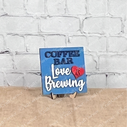 A small blue sign from Janet’s Craft Corner’s Coffee Bar Swappable Squares collection sits on a tan surface against a white brick wall, reading "COFFEE BAR Love IS Brewing" with "IS" inside a red heart—perfect for cozy DIY home decor.