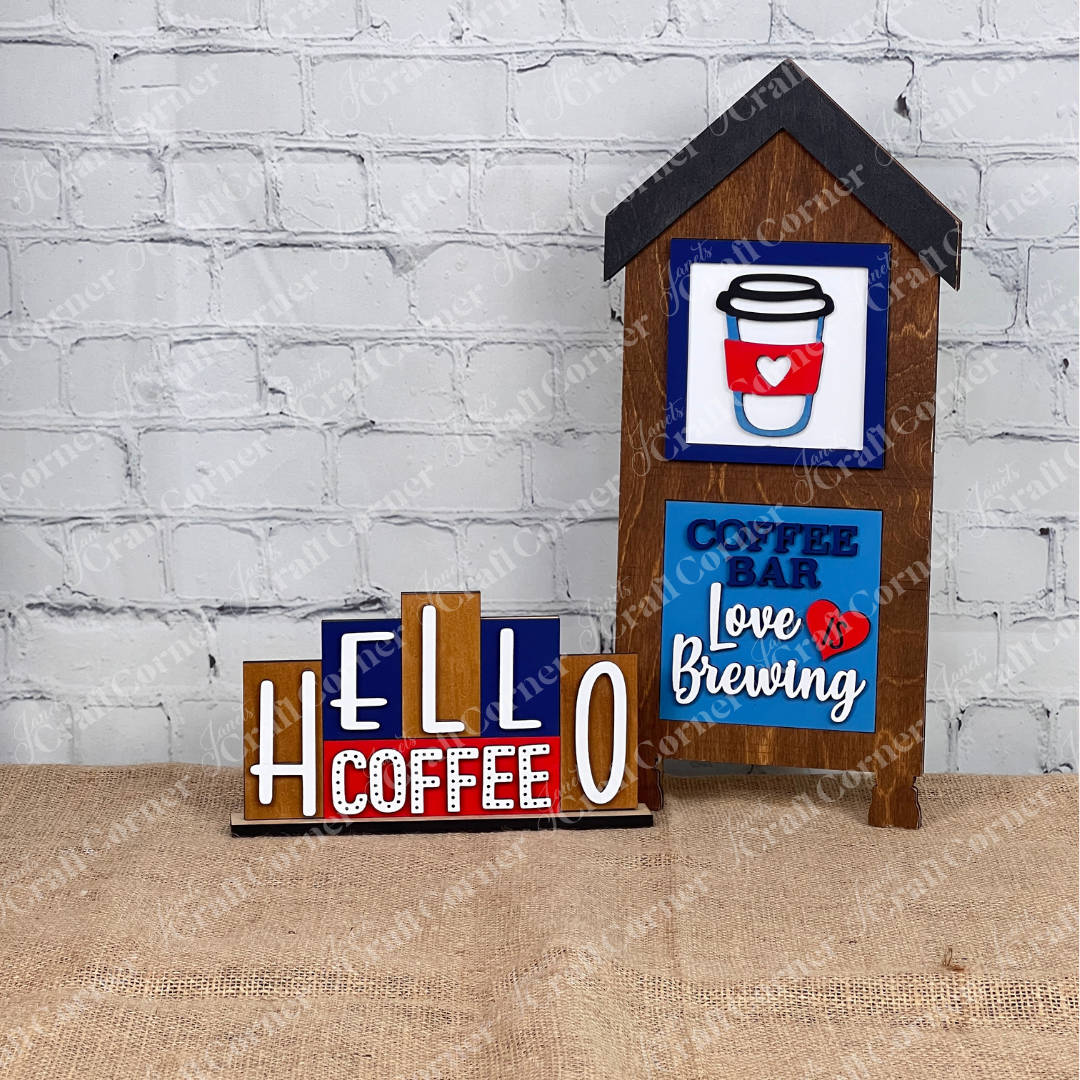 Two “Coffee Bar Swappable Squares” wooden signs by Janet's Craft Corner, perfect for cozy DIY decor, sit in a house shaped frame on burlap, one reads "COFFEE BAR Love is Brewing" with a coffee cup graphic.. A mini word block that says "HELLO COFFEE" sits beside them.