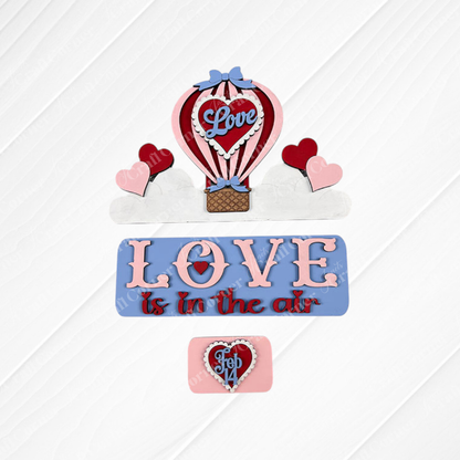 Experience the charm of Janet's Craft Corner with the "Love is in the Air Interchangeable Insert." This DIY home decor kit features decorative cake toppers on a textured surface, showcasing a hot air balloon labeled "Love," hearts, and pastel-colored phrases like "Love is in the Air" and "Feb 14" for an irresistible decorative accent.