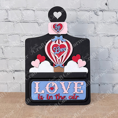 The Breadboard Interchangeable Base with Insert from Janet's Craft Corner is a DIY home decor kit featuring a charming hot air balloon design with "Love" and hearts. "Love is in the air" complements the cozy burlap backdrop against a white brick wall—perfect for creative home decorators.