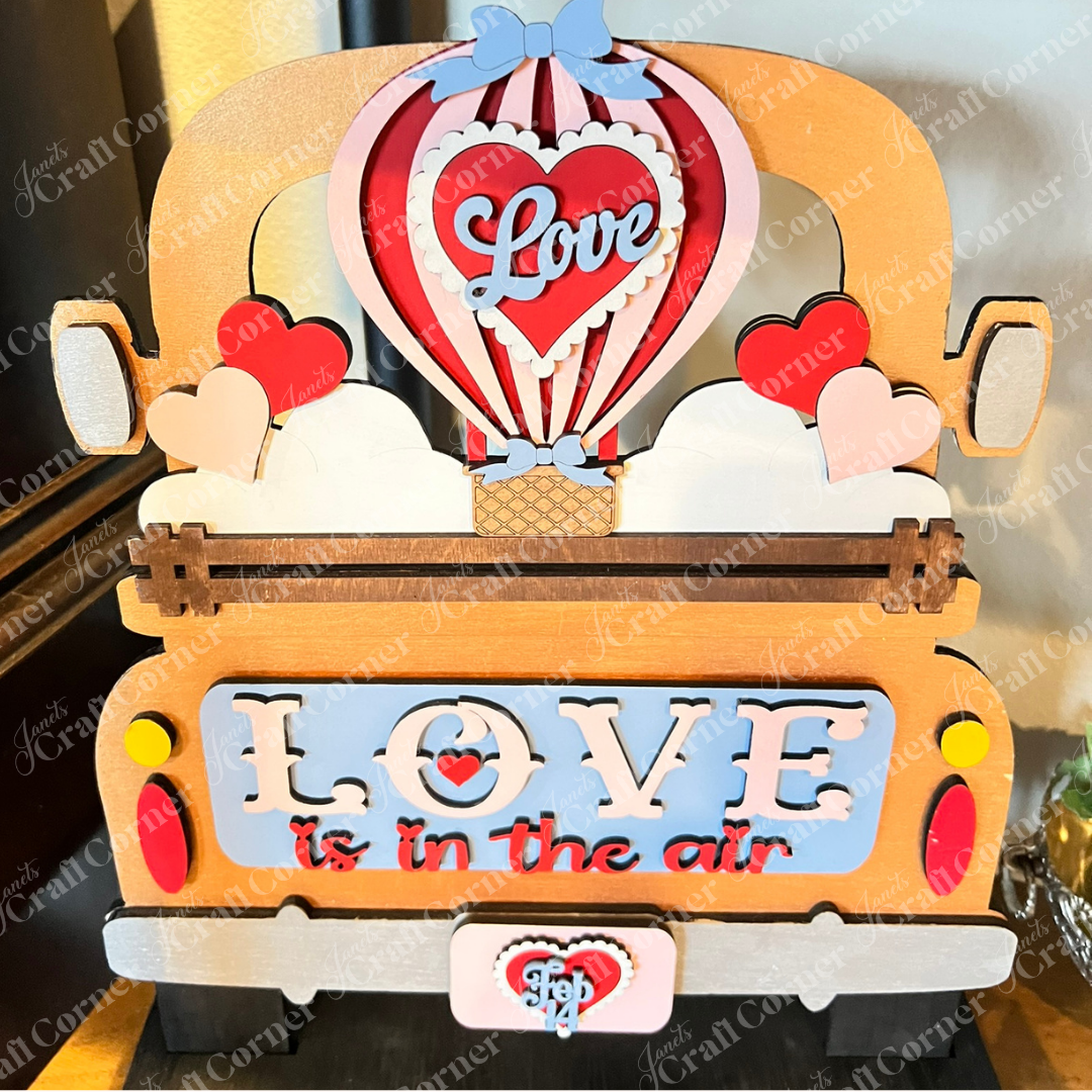 The "Love is in the Air" interchangeable insert by Janet's Craft Corner features a whimsical vintage truck with colorful hearts, a hot air balloon with "Love," and the phrase below. A decorative license plate reads "Feb 14" This DIY décor craft kit includes 3 charming pieces radiating warmth and affection.