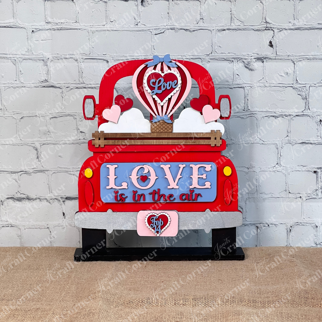 The "Love is in the Air" Interchangeable Insert by Janet's Craft Corner features a decorative sign with a red truck and hearts, "LOVE" on a blue panel, and a hot air balloon labeled "Love" rising from the truck bed against a brick wall backdrop. This DIY kit includes 1 set of 3 pieces.