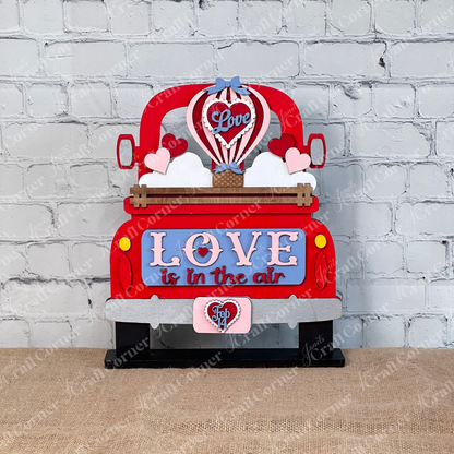 The "Love is in the Air" Interchangeable Insert by Janet's Craft Corner features a decorative sign with a red truck and hearts, "LOVE" on a blue panel, and a hot air balloon labeled "Love" rising from the truck bed against a brick wall backdrop. This DIY kit includes 1 set of 3 pieces.