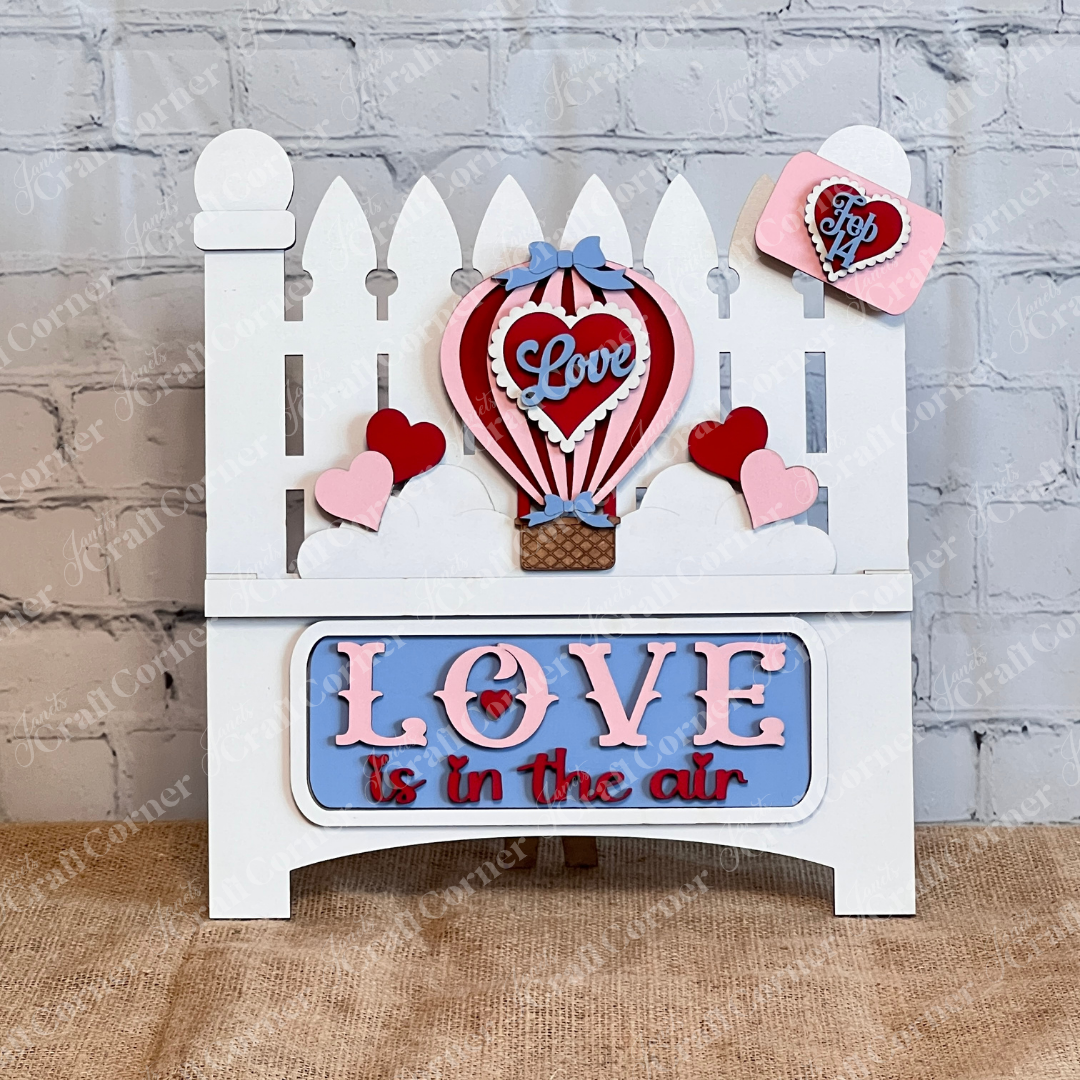 A display features a white picket fence with red and pink hearts, a whimsical hot air balloon labeled "Love," and a blue sign that reads, "Love is in the Air." This captures romance using Janet's Craft Corner's "Love is in the Air" Interchangeable Insert DIY decor kit.