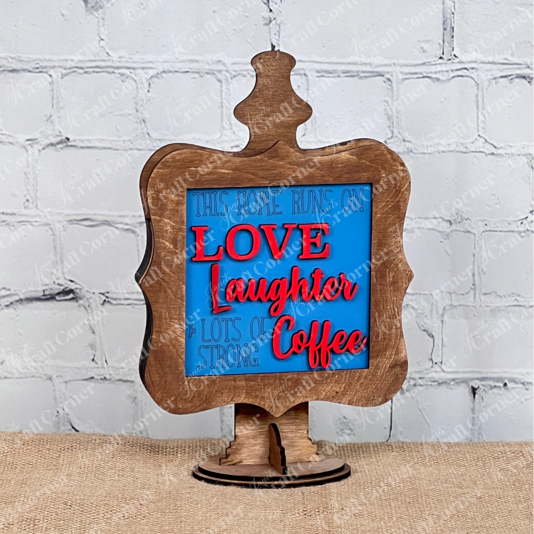 Janet's Craft Corner presents Coffee Bar Swappable Squares—a decorative wooden sign with a blue and red message, "This home runs on love, laughter & lots of strong coffee" displayed in a stained drop-in holder. Ideal for DIY home decor on burlap with a white brick background