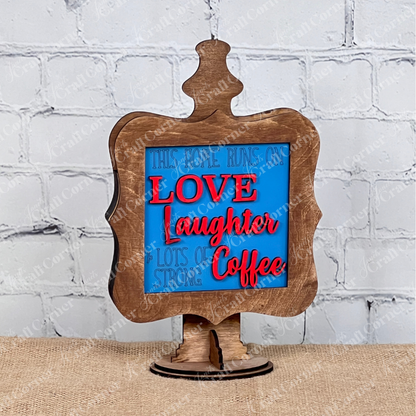 Janet's Craft Corner presents Coffee Bar Swappable Squares—a decorative wooden sign with a blue and red message, "This home runs on love, laughter & lots of strong coffee" displayed in a stained drop-in holder. Ideal for DIY home decor on burlap with a white brick background