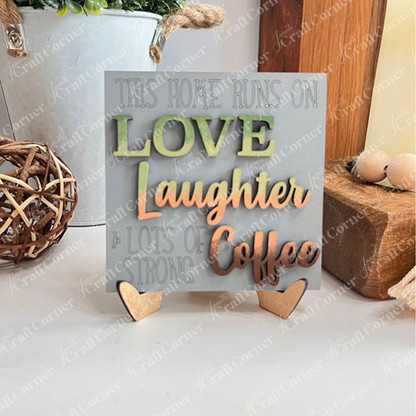 Janet's Craft Corner presents Coffee Bar Swappable Squares—a decorative wooden sign with a green & brown message, "This home runs on love, laughter & lots of strong coffee" displayed on a mini easel. Ideal for DIY home decor on burlap with a white brick background