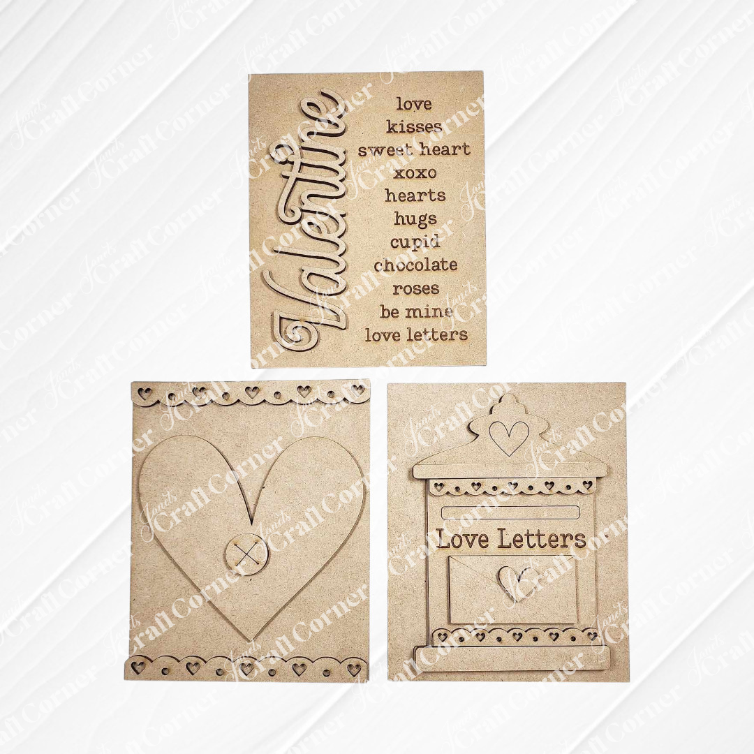 The Love Letters Interchangeable Signs DIY craft kits from Janet's Craft Corner features three charming wooden signs: a country style heart, love-themed words "Valentine," and a mailbox labeled "Love Letters." Set against a white wooden backdrop, they're  perfect for crafting interchangeable home decor.
