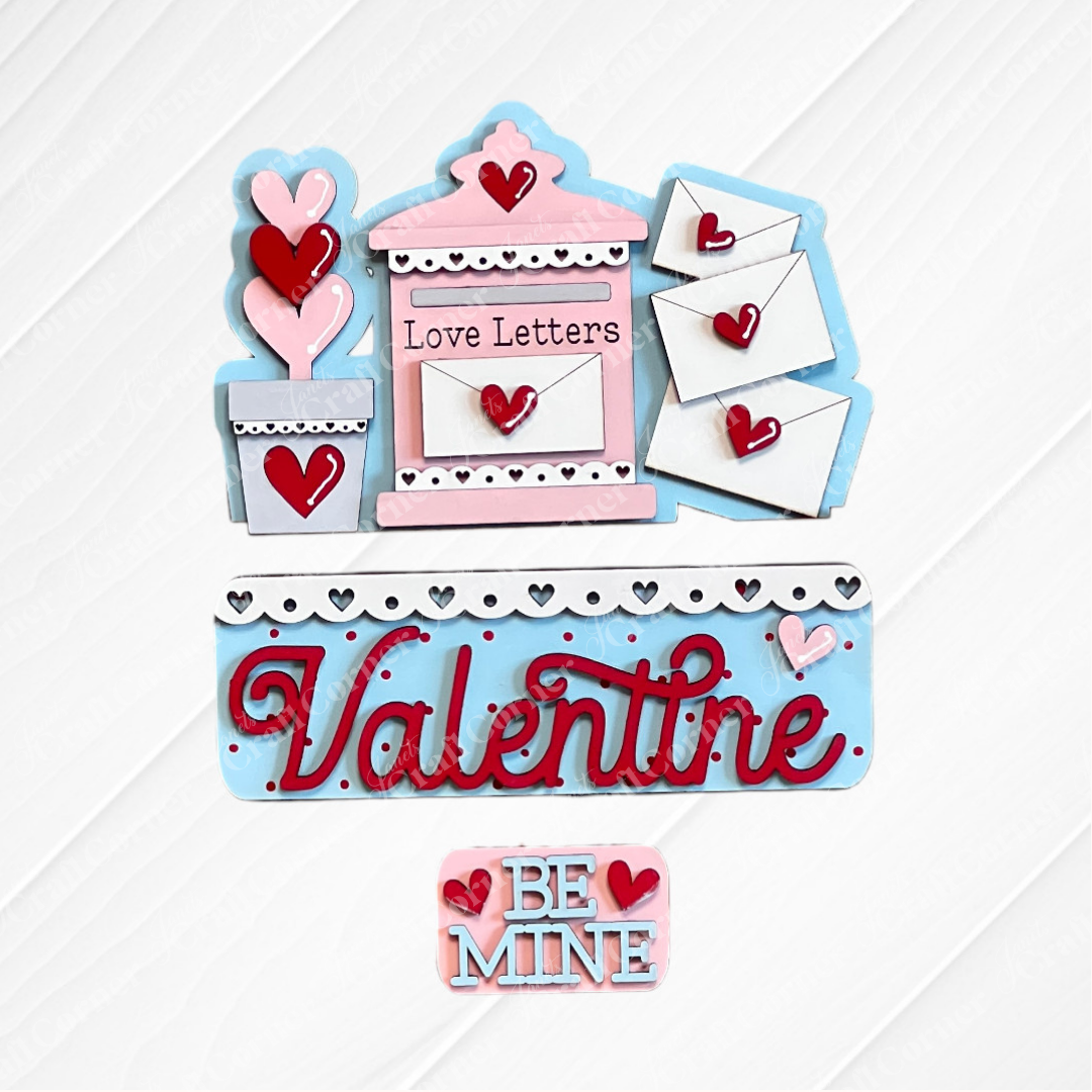 Janet's Craft Corner offers the Love Letters Interchangeable Insert: a Valentine's craft set featuring a pink and blue mailbox labeled "Love Letters," three envelopes with heart seals, and wooden heart cutouts in a flower pot adorned with "Valentine" and "Be Mine" on a white background.