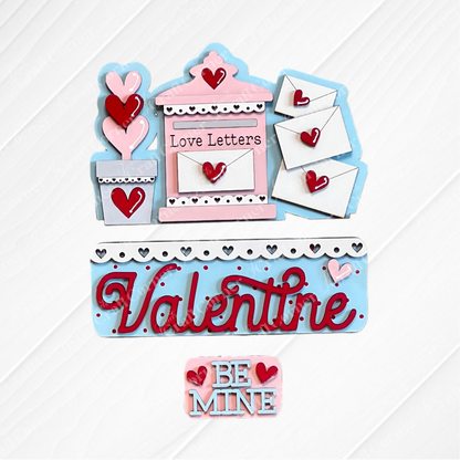 Janet's Craft Corner offers the Love Letters Interchangeable Insert: a Valentine's craft set featuring a pink and blue mailbox labeled "Love Letters," three envelopes with heart seals, and wooden heart cutouts in a flower pot adorned with "Valentine" and "Be Mine" on a white background.