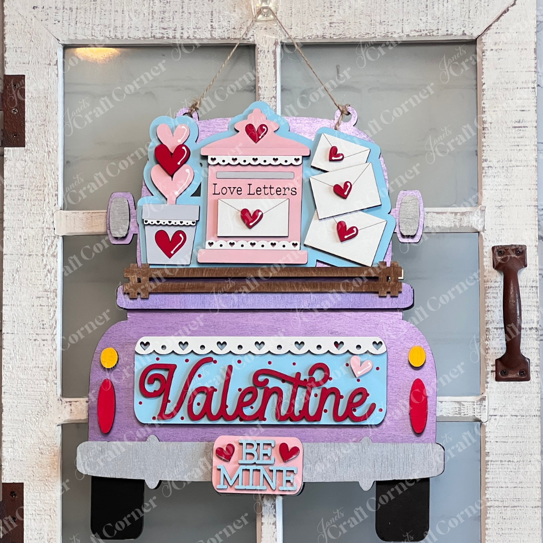 The "Love Letters Interchangeable Insert" by Janet's Craft Corner is a decorative wooden sign shaped like a truck, with "Valentine" on it and a "Be Mine" sign below. Set against a white brick wall and window, it's perfect for adding Valentine's Day charm to your home.