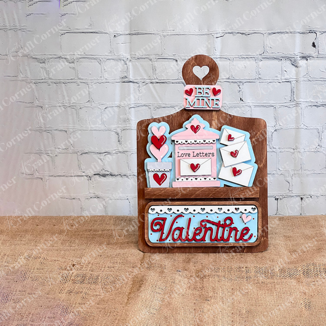 Janet's Craft Corner offers the Love Letters Interchangeable Insert DIY kit, featuring Valentine's decor with red and pink hearts, a "Be Mine" sign, and a customizable envelope design. The word "Valentine" in red and blue stands out against a brick wall backdrop.