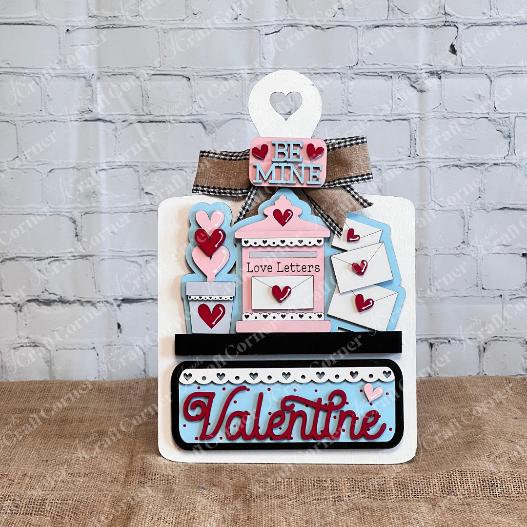 Janet's Craft Corner offers a DIY Valentine's Day decor kit with a breadboard base and insert. It features phrases like "Be Mine" and "Love Letters," adorned with hearts. With pink, blue, and red accents, it enhances home decor beautifully against a brick wall background.