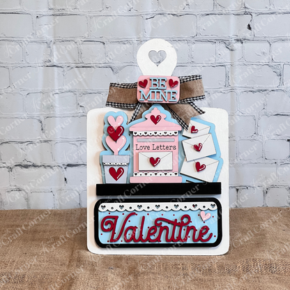 Janet's Craft Corner offers a DIY Valentine's Day decor kit with a breadboard base and insert. It features phrases like "Be Mine" and "Love Letters," adorned with hearts. With pink, blue, and red accents, it enhances home decor beautifully against a brick wall background.