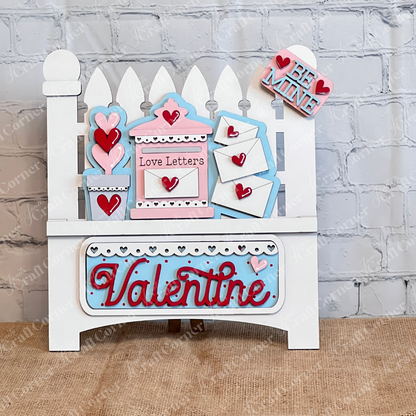 The Love Letters Interchangeable Insert from Janet's Craft Corner is a decorative wooden piece featuring Valentines-themed items like hearts and envelopes with pastel words "Love Letters" and "Valentine," set against a white picket fence in front of a white brick wall.