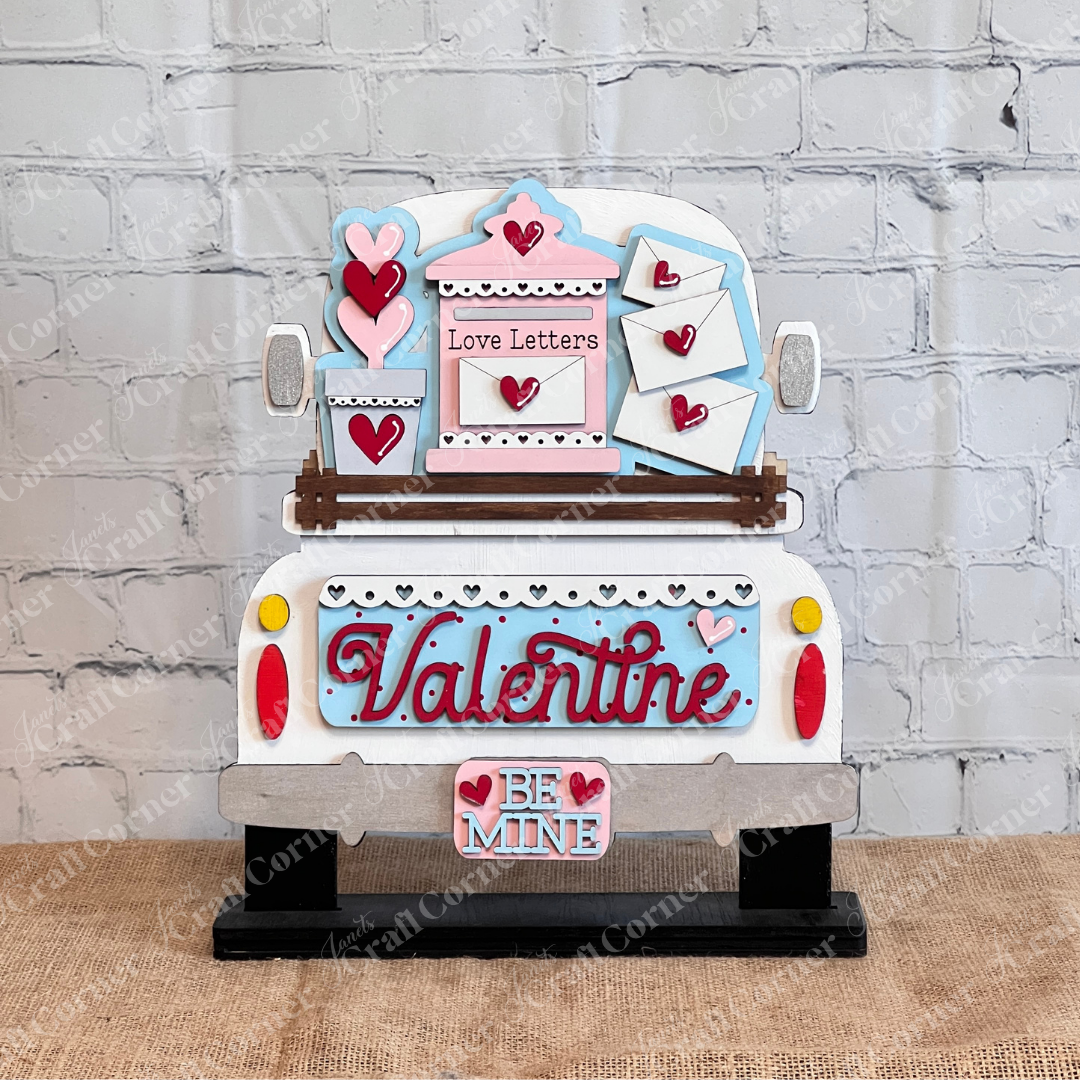 A decorative display features a vintage truck with "Valentine" and "Be Mine," red hearts, envelopes, and a "Love Letters" sign. Set against a white brick background on burlap, this is part of the Love Letters Interchangeable Insert by Janet's Craft Corner for personalized Valentine's Day decor.