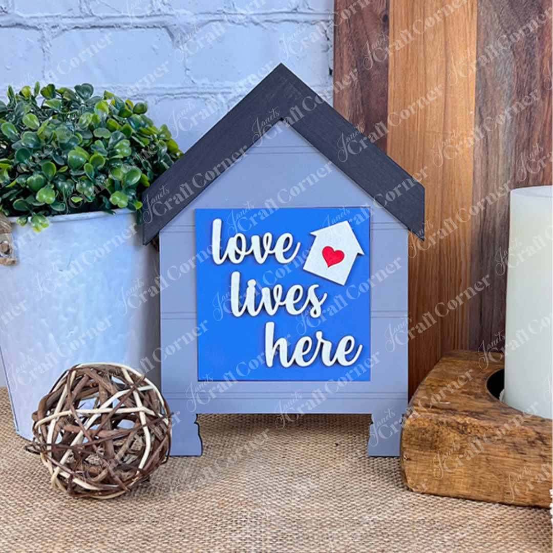 The Home Squares sign by Janet's Craft Corner features a small, house-shaped design with a black roof, the phrase “love lives here,” and a red heart—perfect tiered tray decor. Shown with a plant, twig ball, and wooden block.