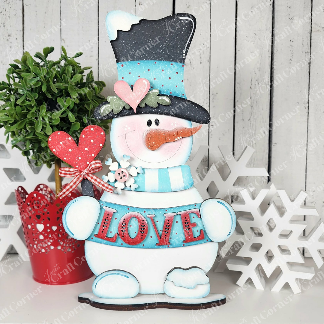 The Love Snowman Shelf Sitter by Janet's Craft Corner features a smiling snowman with a black hat, blue scarf, and red heart holding a "LOVE" sign. DIY kit includes shelf sitter, red potted plant, and snowflake decorations for seasonal decor.