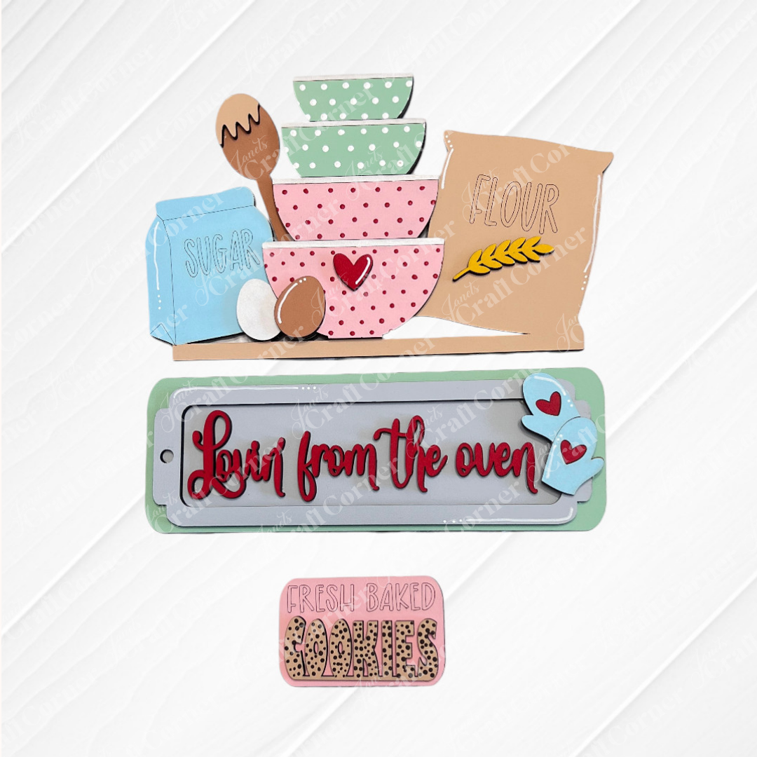 The Lovin from the Oven Interchangeable Insert by Janet's Craft Corner is a DIY home décor craft kit featuring wall hangings of stacked pastel bowls, a spoon, eggs, sugar and flour bags. It includes signs saying "Love from the oven" and "FRESH BAKED COOKIES," with a textured burlap background for rustic charm.
