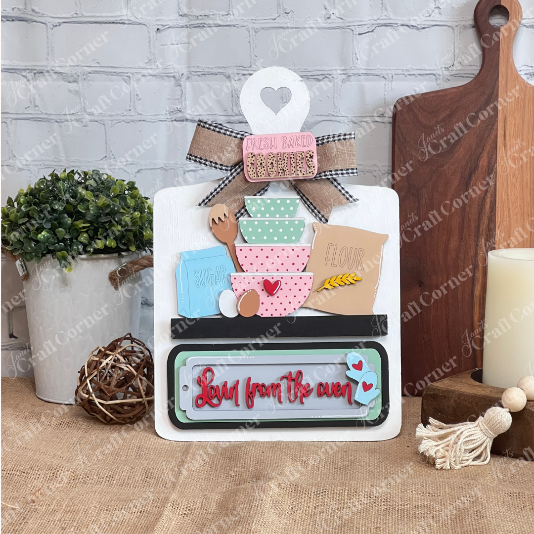 The Lovin from the Oven Interchangeable Insert by Janet's Craft Corner is a DIY home décor craft kit featuring stacked bowls, a rolling pin, and boxes labeled "Flour" and "Sugar" against a brick wall. It includes a ribbon on top and "Love from the oven" in red script at the bottom. Perfect for DIY projects.