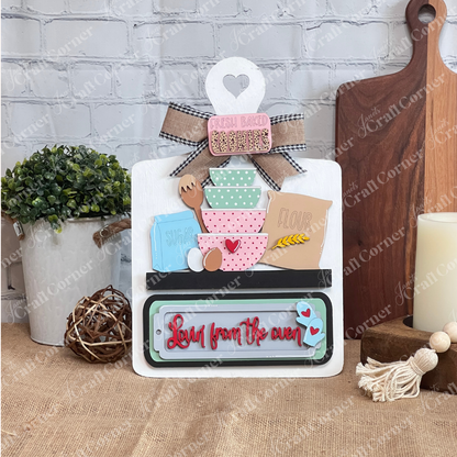 The Lovin from the Oven Interchangeable Insert by Janet's Craft Corner is a DIY home décor craft kit featuring stacked bowls, a rolling pin, and boxes labeled "Flour" and "Sugar" against a brick wall. It includes a ribbon on top and "Love from the oven" in red script at the bottom. Perfect for DIY projects.