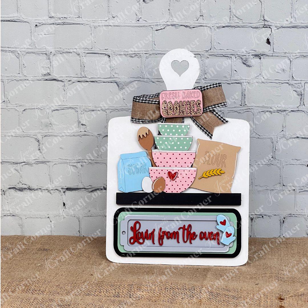 Add charm to your home decor with Janet's Craft Corner Breadboard Interchangeable Base and Insert. The DIY kit includes a breadboard base and an insert featuring pastel bowls labeled with baking essentials, topped by "Fresh baked cookies," and "Love from the oven" set against a burlap backdrop.