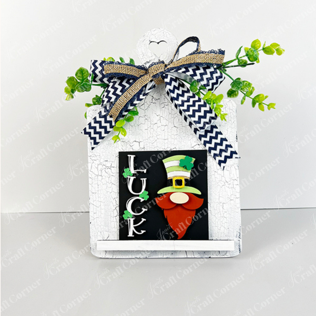 Janet's Craft Corner's Lucky Irish Squares is a house-shaped decorative sign with a green-hatted gnome, "LUCK" wood cut-outs, a chevron bow with greenery, and cracked white paint—ideal for tiered tray displays.