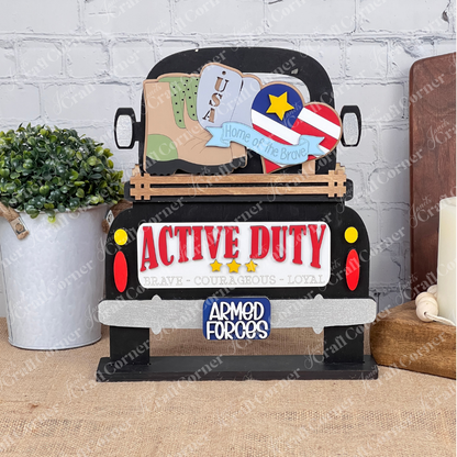 A charming truck-shaped sign labeled "Active Duty," adorned with military service symbols like boots, a star, and a heart inscribed with "Home of the Brave," is part of Janet's Craft Corner's Military Interchangeable Insert DIY home decor kit. The display also includes a small plant and candle on the table.