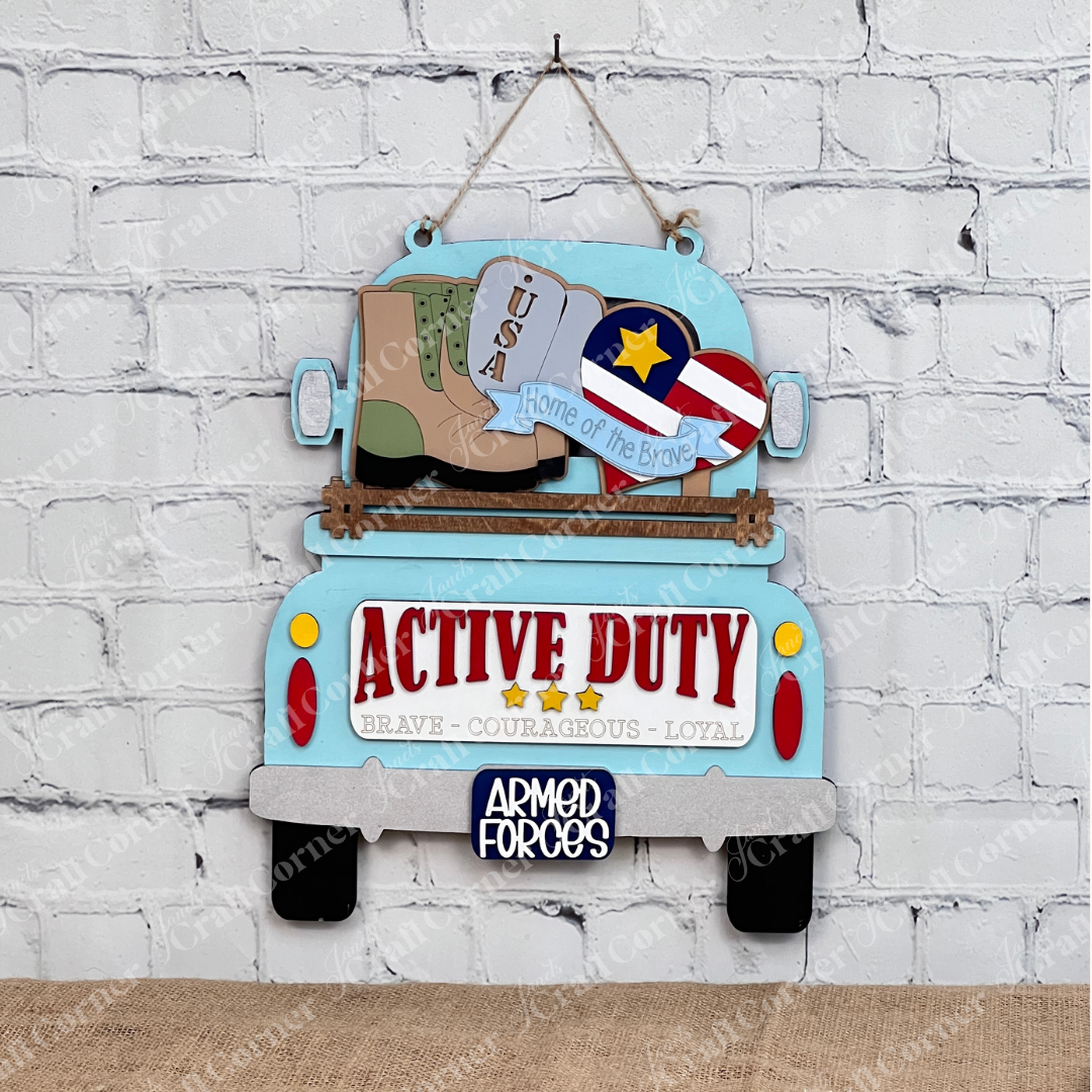 Janet's Craft Corner offers a Military Interchangeable Insert DIY kit featuring a blue vintage truck sign with "Active Duty" on the tailgate, military boots, USA dog tags, and a star emblem. Ideal for home decor against a white brick wall.