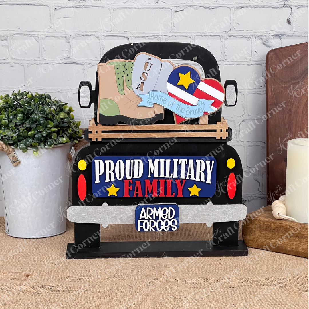 The Military Interchangeable Insert by Janet's Craft Corner is a decorative sign that showcases a vintage truck adorned with the phrases "Proud Military Family" and "Armed Forces." The truck bed features a boot, USA sign, and heart with flag design. This DIY home decor kit comes equipped with elements like a potted plant, candle, and brick wall to enhance your patriotic display at home.