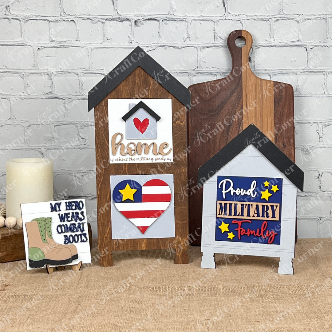 Janet's Craft Corner displays patriotic wood cut-outs, including house-shaped "home" signs, a stars-and-stripes heart, "Proud Military Family," and a Military Squares sign that reads "My Hero Wears Combat Boots," all against a white brick wall.