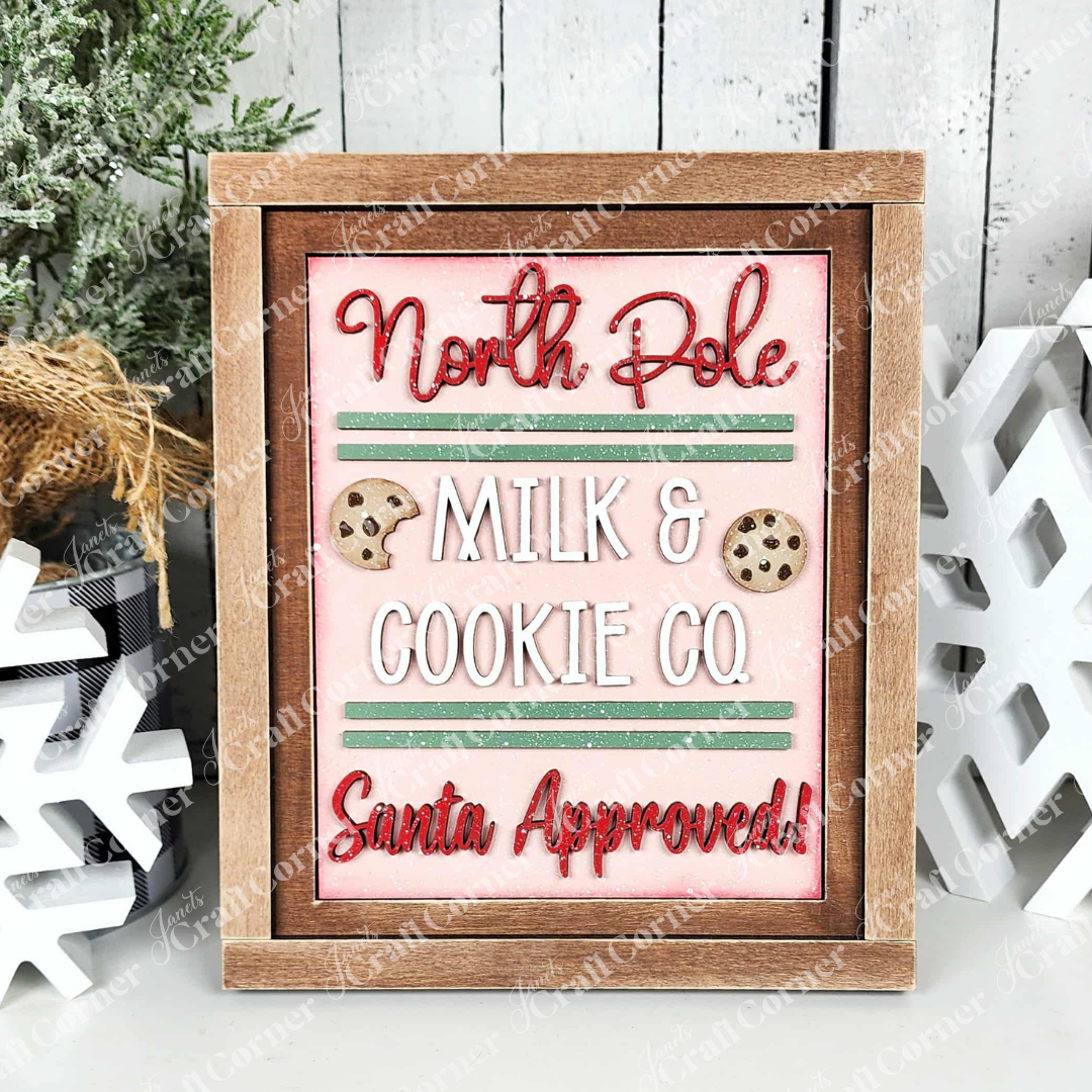 The North Pole Switchable Farmhouse Signs from Janet’s Craft Corner features festive red and green text, cookie illustrations, and green stripes, perfect for DIY seasonal decor with tree branches and snowflakes.