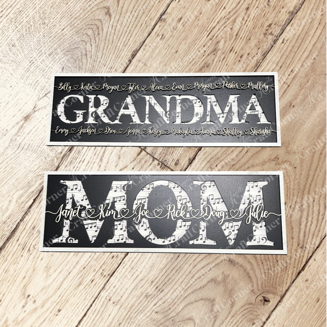 Two rectangular black Personalized Mom Signs from Janet's Craft Corner sit on a wooden floor—one says "GRANDMA" with smaller names, the other "MOM" with several cursive names—perfect custom gifts for moms or grandmas.