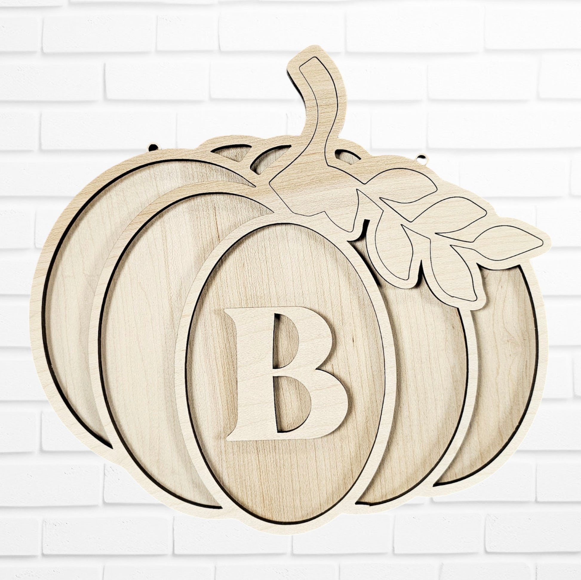 Janet's Craft Corner presents the Monogram Pumpkin Wall Hanger, a DIY home decor craft kit featuring a wooden cutout shaped like a pumpkin with a prominent letter "B" in the center and detailed leaf accents at the stem. It's elegantly displayed against a white brick wall for perfect seasonal charm.