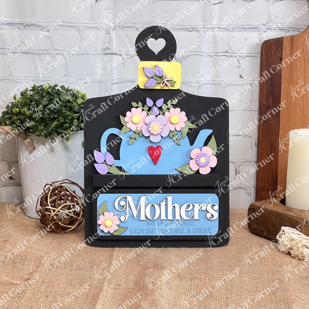 Janet's Craft Corner offers a DIY interchangeable home decor kit featuring a breadboard base and an insert with a teapot brimming with pink flowers. The accompanying message, "Mothers are like flowers, each one beautiful & unique," is adorned with heart and flower motifs. Perfect for personalization.