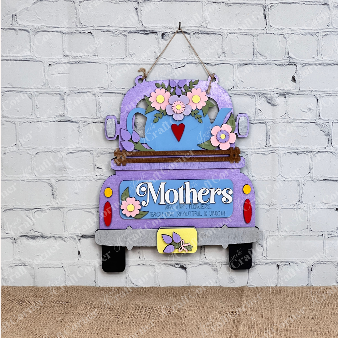 A delightful display piece from Janet's Craft Corner, the "Mothers Interchangeable Insert - Painted" on a decorative sign shaped like a purple vintage truck. It features a charming teapot motif adorned with colorful flowers on its roof, against which the phrase "Mothers are like flowers, each one beautiful & unique" is elegantly displayed. The sign sits gracefully on a wooden surface with a brick wall providing the perfect backdrop.