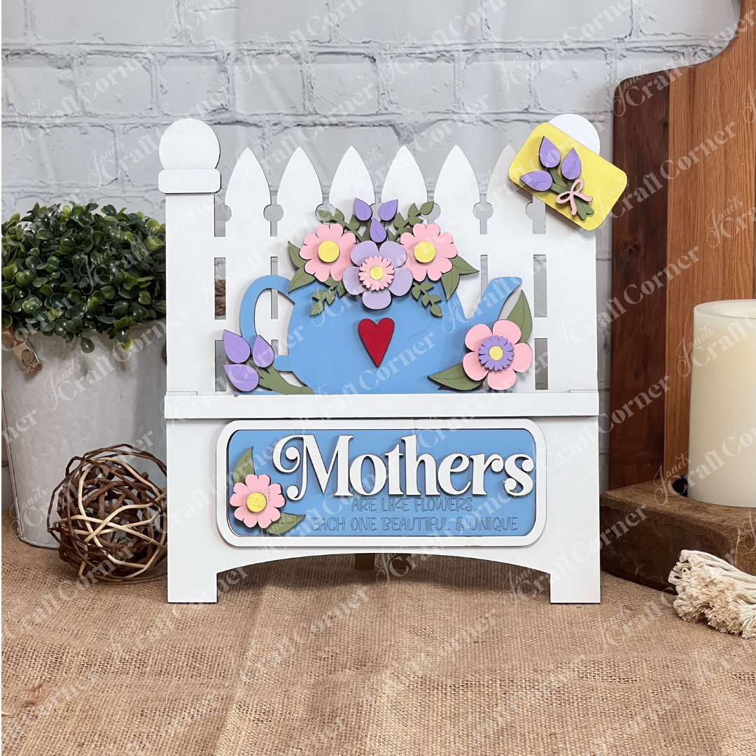 The Garden Fence with Insert by Janet's Craft Corner features a picket fence design adorned with a blue teapot and flowers, bearing the phrase "Mothers are like flowers, each one beautiful & unique." It's the perfect way to celebrate maternal love against a white brick wall.
