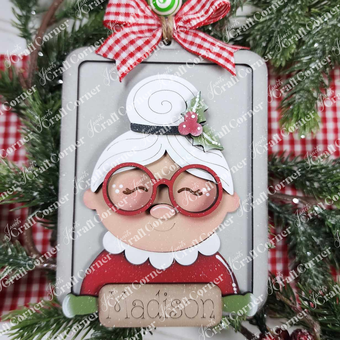 Add festive charm with Janet's Craft Corner's Mrs. Claus Ornament, featuring a cartoon grandmother in a red "Madison" outfit, glasses, white hair bun, and finished with a red plaid bow and holly—perfect for Santa ornament lovers.