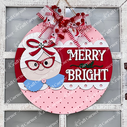 Janet's Craft Corner Mrs. Claus Merry & Bright Round Sign DIY craft kit includes a festive door sign with Mrs. Claus, ribbons, polka dots, and "Merry & Bright" in white and green on a red and pink background. Set of 1.