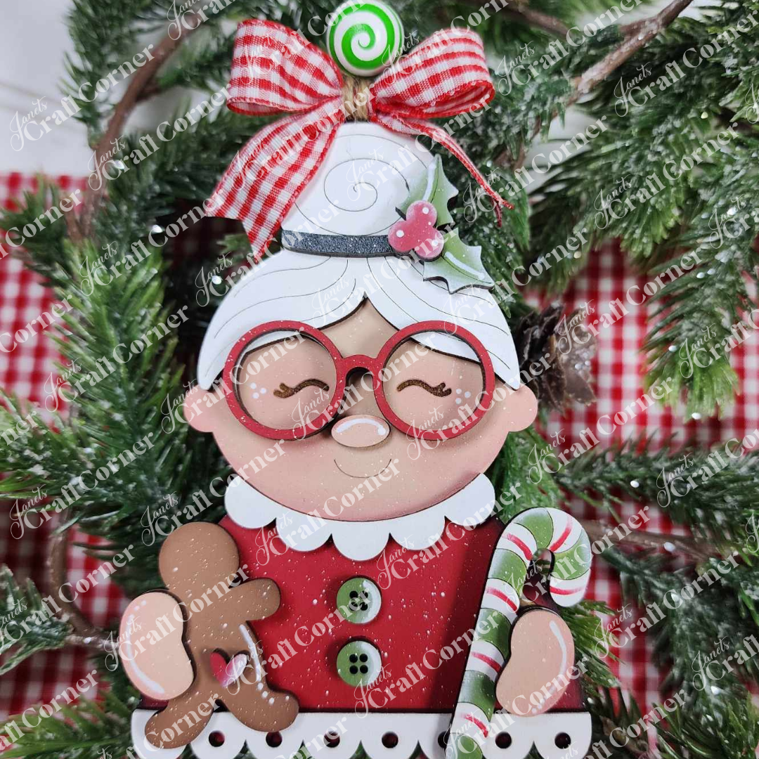 The Mrs. Claus Ornaments by Janet's Craft Corner feature Mrs. Claus with red glasses, a red dress, and white hair, holding a gingerbread cookie and candy cane, hanging on a Christmas tree with a red plaid bow.