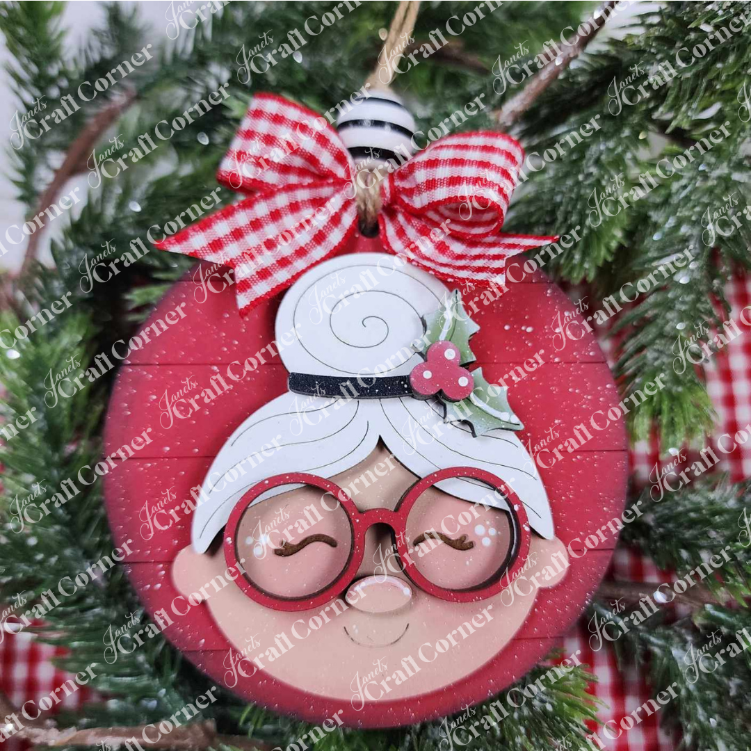 The Mrs. Claus Ornaments by Janet's Craft Corner feature a round red design with Mrs. Claus’ smiling face, glasses, bun, holly leaves, and a gingham bow—perfect for adding charm to your festive tree décor.