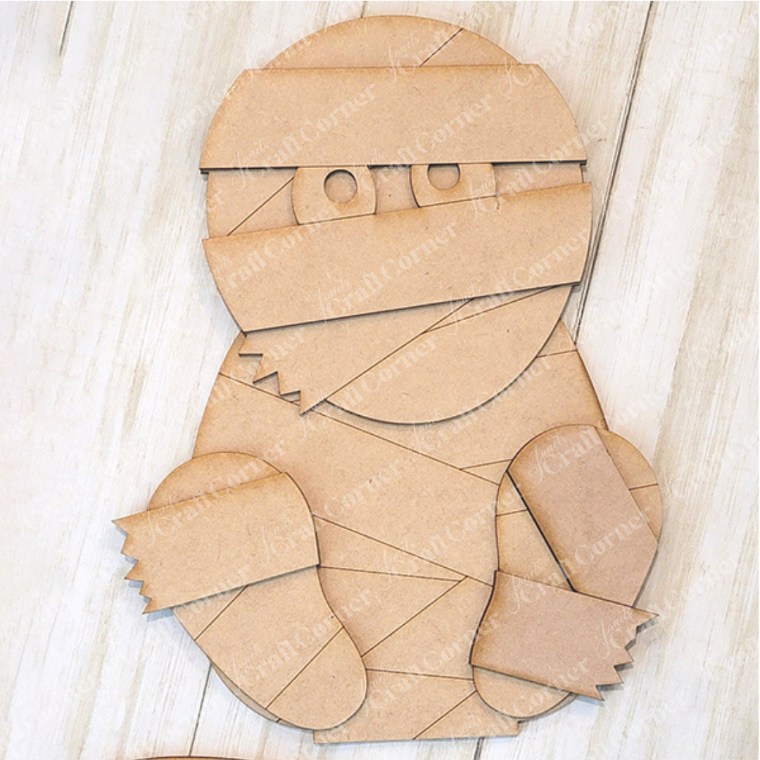 This delightful mummy shelf sitter from Janet's Craft Corner is a DIY home decor kit made of simple geometric shapes—a round head, rectangular body, and triangular limbs—crafted from cardboard. It rests on a light wooden surface, inviting your creative touch to bring it to life.