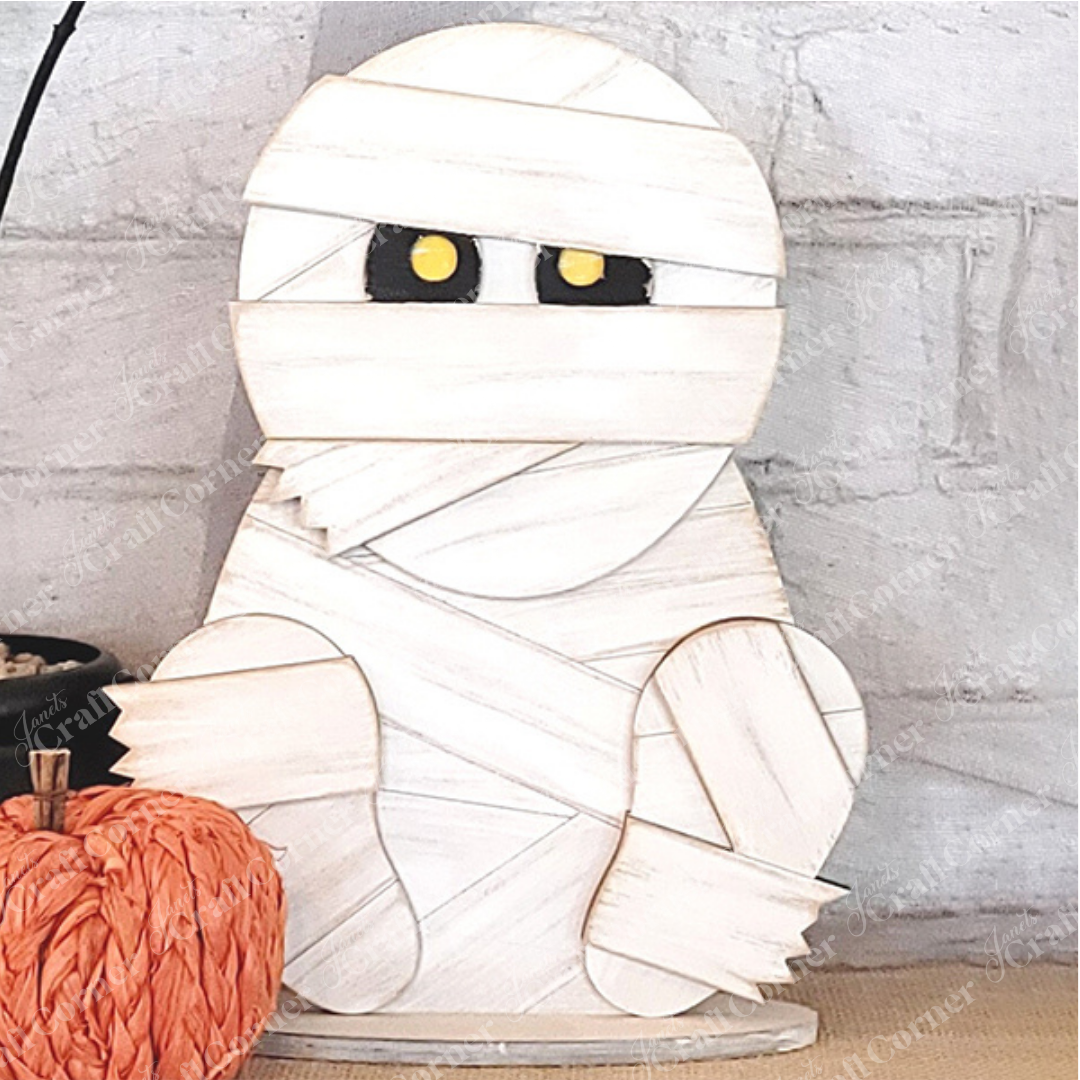 The Mummy Shelf Sitter DIY home decor kit by Janet's Craft Corner features a cartoon-style figure with yellow eyes wrapped in white bandages, perfect for positioning next to a small orange pumpkin. Enthusiasts of hand-painted DIY projects will appreciate using it to create a charming display against a white brick background.