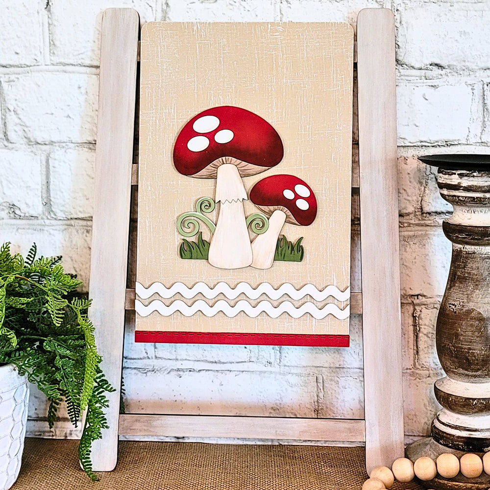 The Mushroom Tea Towel from Janet's Craft Corner, a delightful piece of home décor, showcases two cartoon red and white mushrooms on a beige background. A wooden stand frames the towel beautifully, complemented by green plants and a wooden candlestick against a white brick wall.